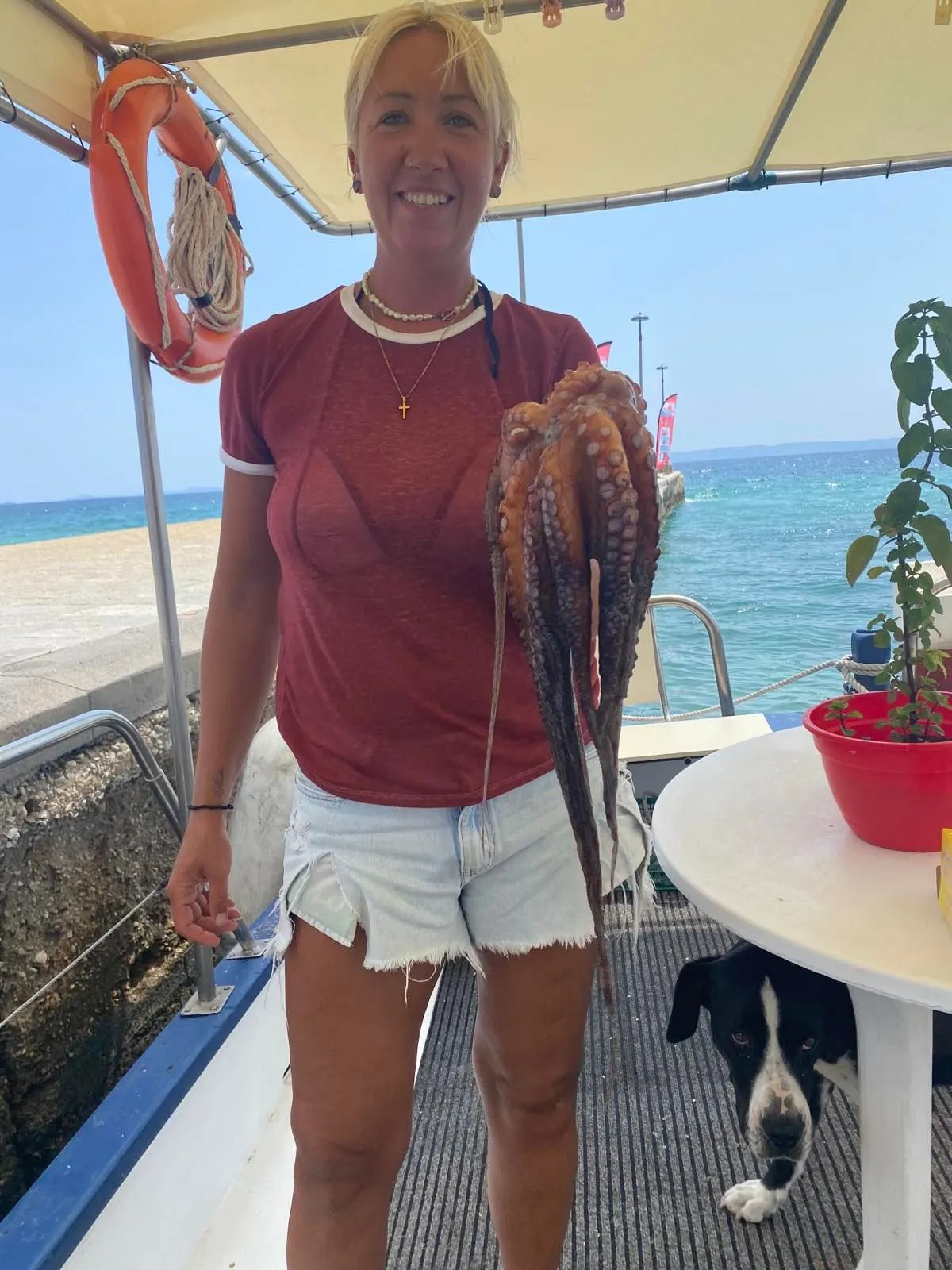 A crew member holding an octopus at the quay, with one of the boat dogs beside her